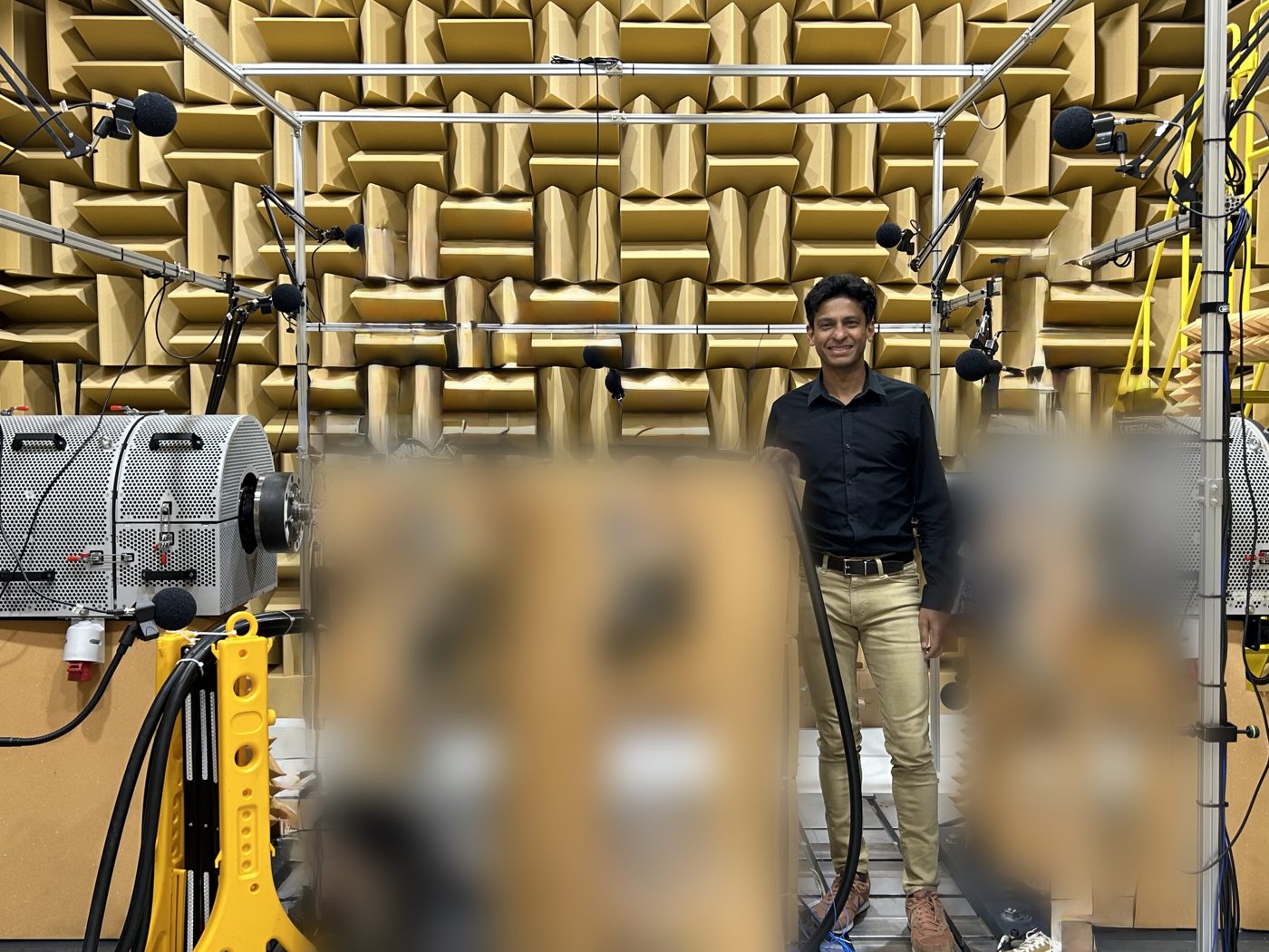 Hasan Pasha standing inside an anechoic chamber lined with sound-absorbing wedges, microphones on booms around a test stand. The test hardware is blurred.